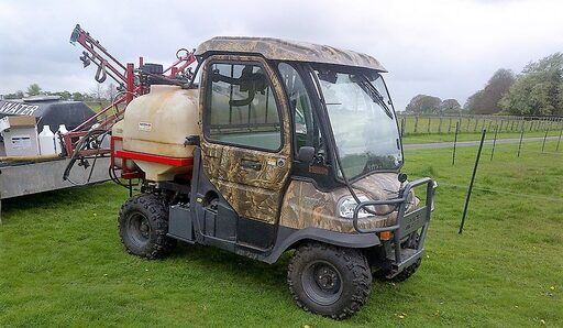 Camouflage utility vehicle with mounted sprayer equipment parked on grass near fields and fencing.